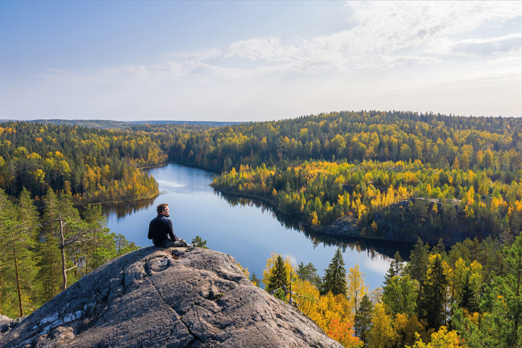 A person sitting on top of a tall rock in an autumn forest. There is a lake in the background.