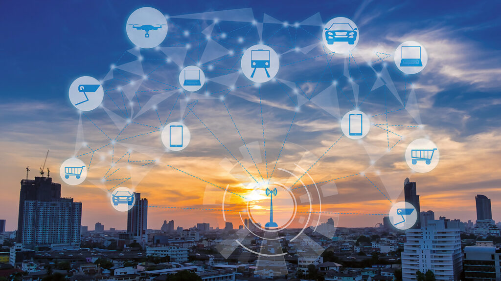 A city at sunset. There are different icons in the front that represent cars, laptops, phones, shopping carts, and a drone. All icons are connected to each other and a single icon of an antenna.