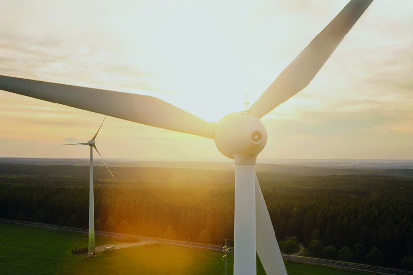 Two wind turbines photographed from the air. Forest and sunset in the background.