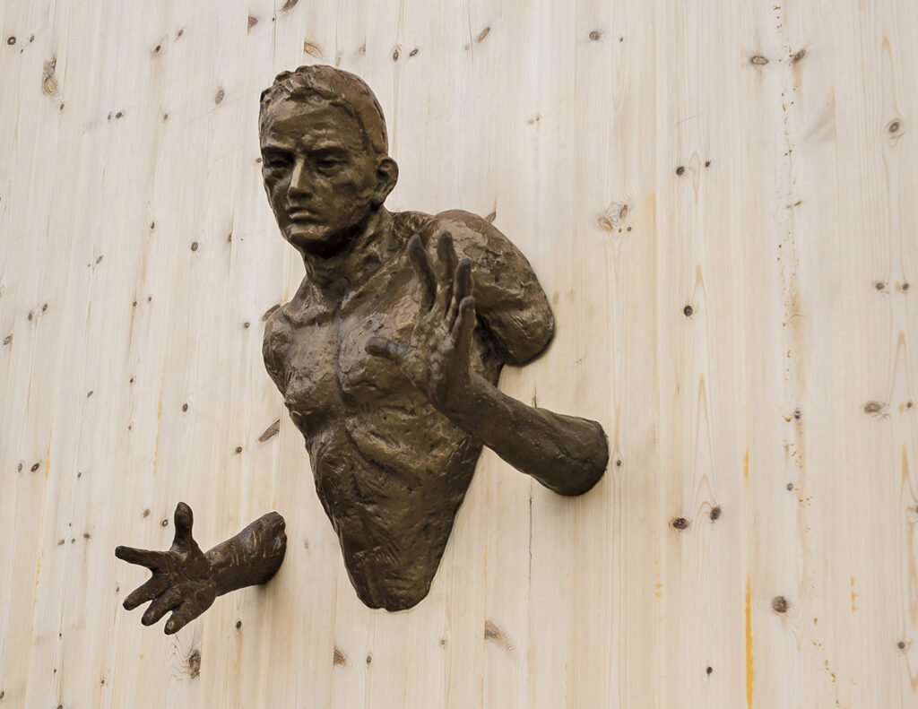 A human statue mounted on a wooden wall. Only parts of the body are visible, making it look like the statue is coming through the wall.