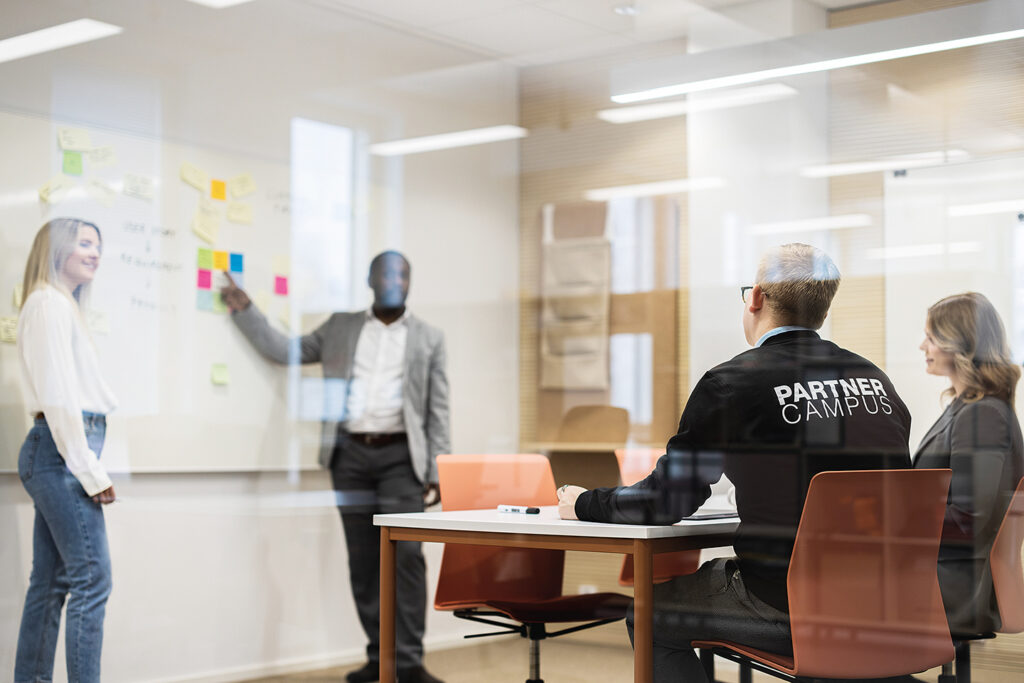 Two people giving a presentation in front of a white board. Two other people are watching the presentation, one of which is wearing a shirt that says “Partner Campus” in the back.