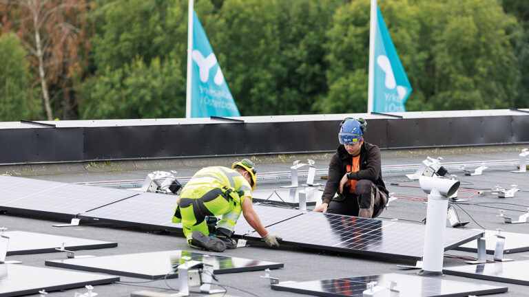 Two people installing a solar panel on a roof. Two flags of Yrkesakademin in the background.