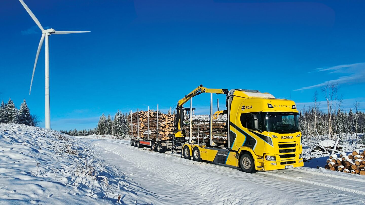 Electric timber truck with a crane and a wood load on a snowy road. There is a wind turbine in the background.