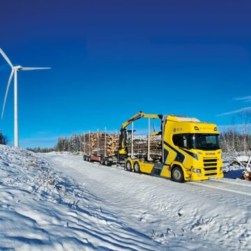 Electric timber truck with a crane and a wood load on a snowy road. There is a wind turbine in the background.