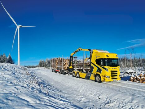 Electric timber truck with a crane and a wood load on a snowy road. There is a wind turbine in the background.