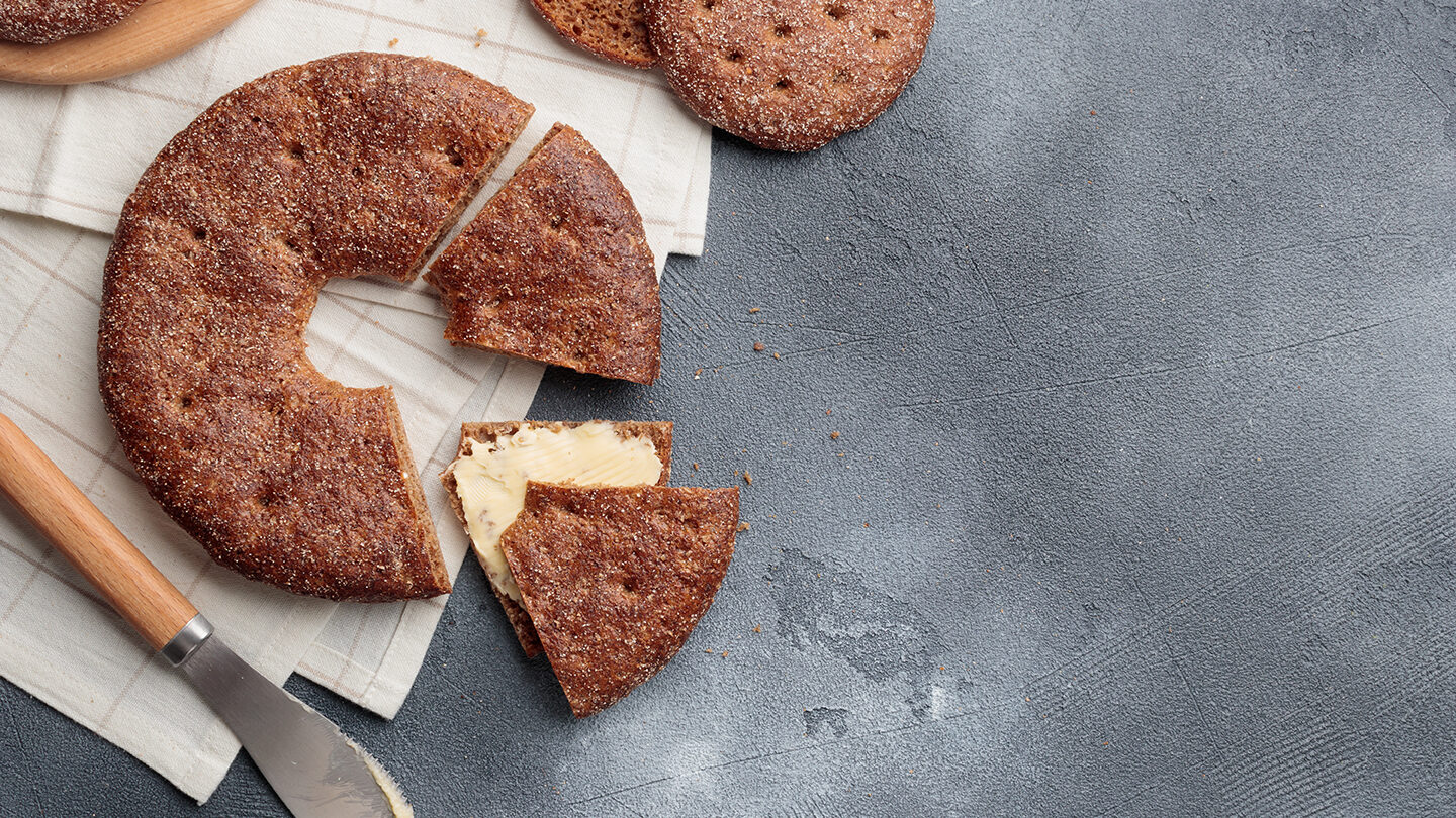 A round rye bread. One piece has been cut from it and butter has been spread on it. There is a butter knife next to the bread.