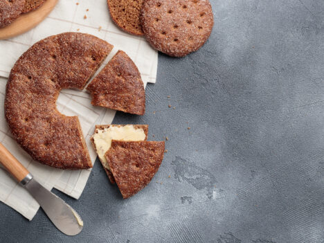 A round rye bread. One piece has been cut from it and butter has been spread on it. There is a butter knife next to the bread.