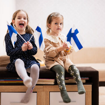 Two small kids sitting on a bench in a living room holding Finnish flags.