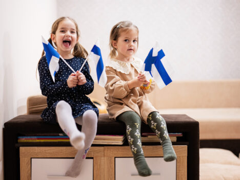 Two small kids sitting on a bench in a living room holding Finnish flags.