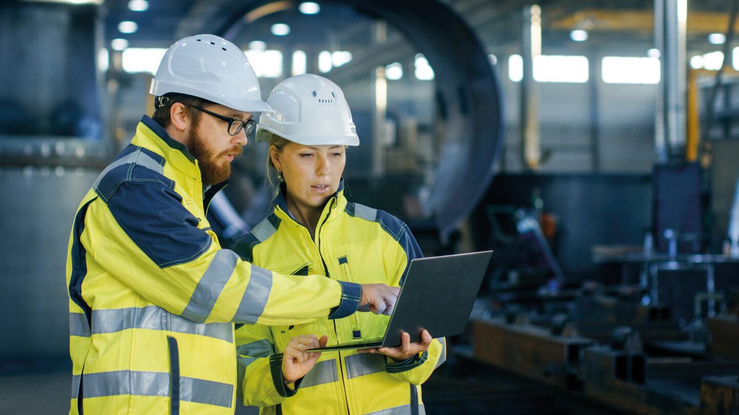 Two people using a laptop. Both are wearing protective workwear and helmets.
