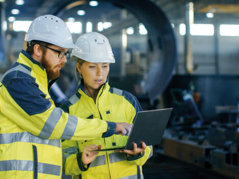 Two people using a laptop. Both are wearing protective workwear and helmets.